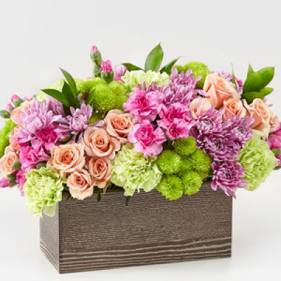 Capture the beauty of the seasons in bloom with our Simple Charm Bouquet. Gorgeous blooms such as peach spray roses, green trick dianthus, pink mini carnations and lavender cushion pompons fill a weathered wooden box with freshness.