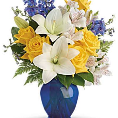 Like a sunny day at the shore, this bright bouquet invigorates and inspires! Radiant yellow roses, white lilies and blue delphinium are expertly arranged in our bold blue plastic ginger jar.
Sunny yellow roses, white asiatic lilies, white alstroemeria, and blue delphinium are arranged with leatherleaf fern and lemon leaf.